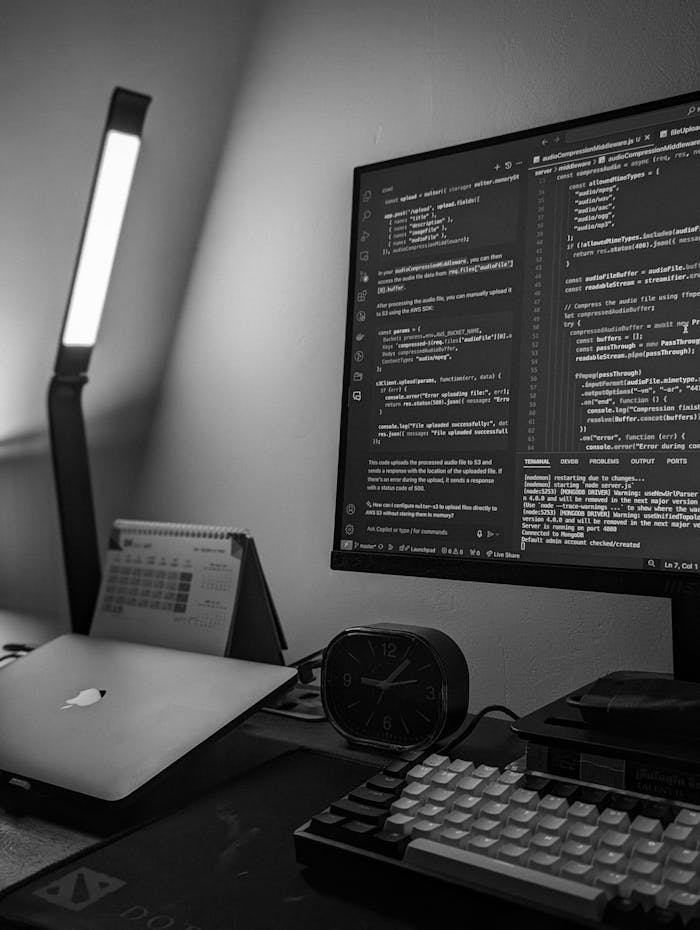 Black and white photo of a workspace featuring a monitor with coding screen, laptop, and desk lamp.